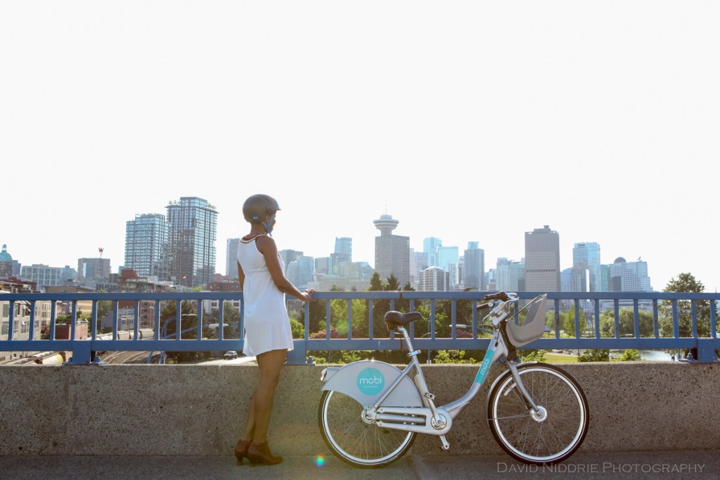 A woman looks out over Vancouver city with a Mobi bike share bicycle by her side.
