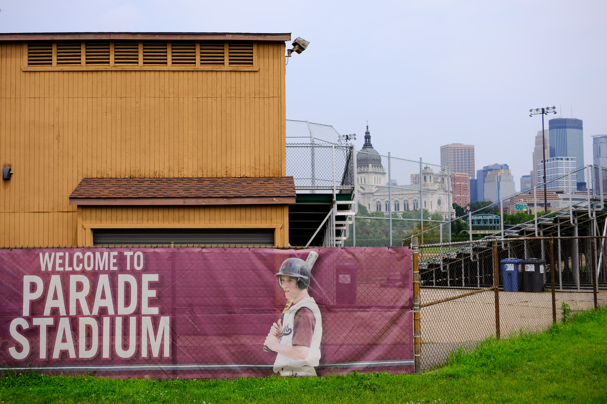 The back of a baseball stadium is shown with a fence featuring a player and a welcome sign with skyline in the background.