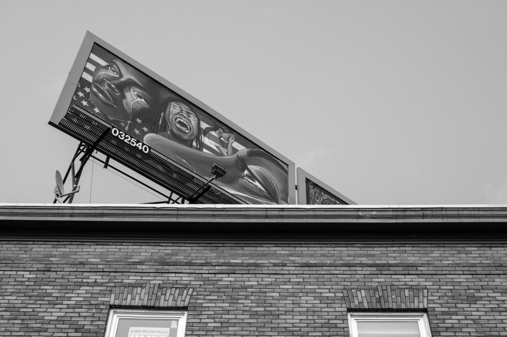 A black and white image shows a billboard on a rooftop with artwork of people crying.
