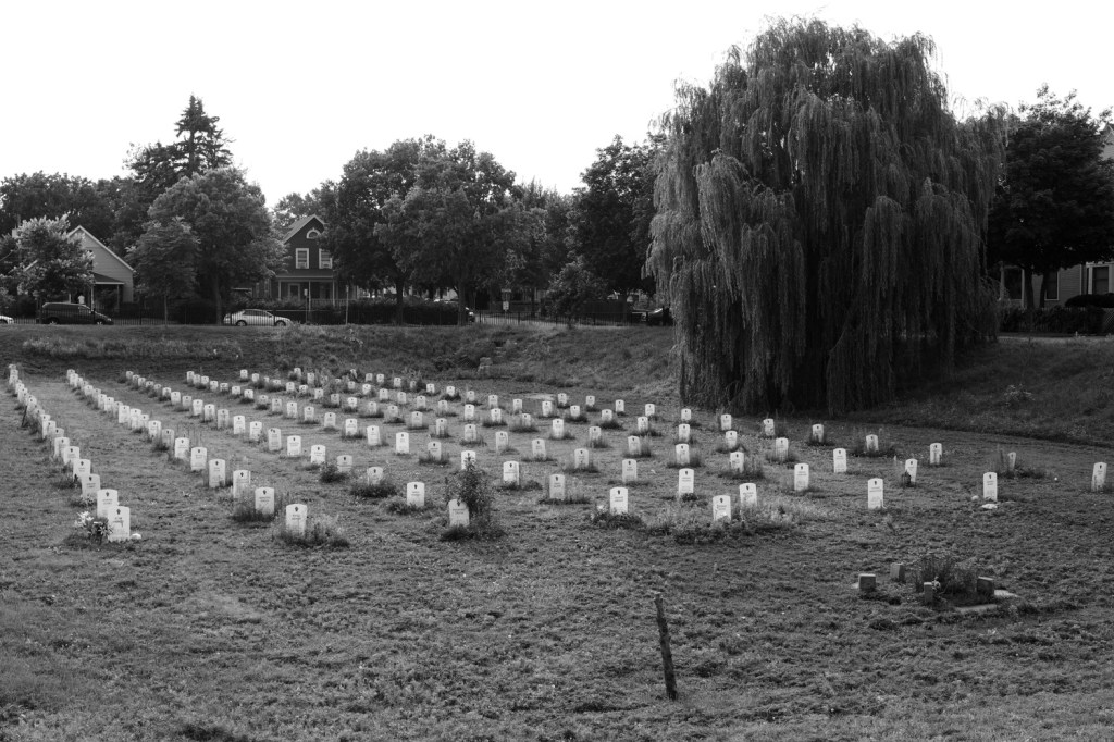 A black and white image shows the Say Their Names Cemetary art installation - 100 white gravestones with the names of black people killed by police. 
